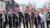 Acht Erwaschene Personen auf einer Baustelle, alle tragen einen roten Helm und haben einen Spaten in der Hand