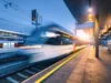 Langzeitbelichtetes Bild eines einfahrenden Zuges im Bahnhof, der durch die Bewegung verschwommen wirkt, mit beleuchteter Bahnsteigumgebung in der Dämmerung.