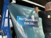 Mitarbeiter bringt eine Fahrzeugbeklebung mit dem Text „Mit Ökostrom zum Ziel“ an einem blauen Bus an.