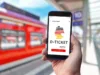 Nahaufnahme einer Hand, die ein Smartphone hält, auf dem das Angebot für das Deutschlandticket exemplarisch abgebildet ist. Im Hintergrund ist ein Zug zu sehen.